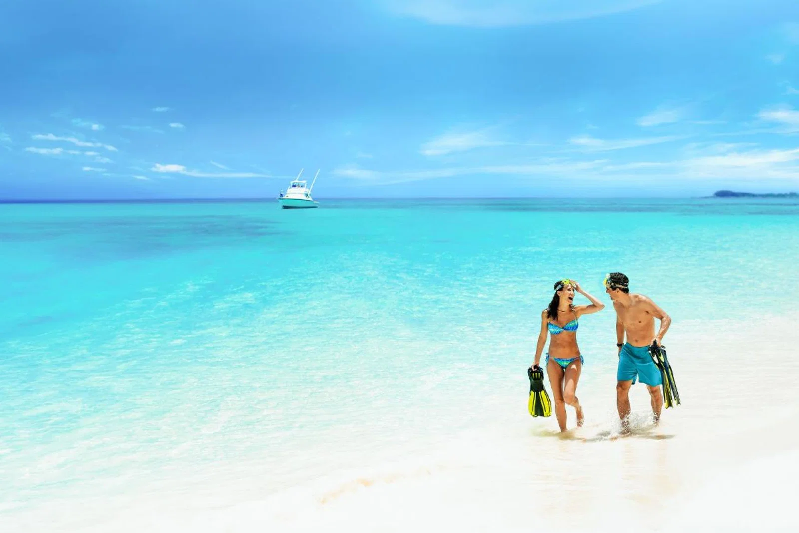 A young couple in swimwear walk along the white sand beach with their feet in the shallow of the blue sea
