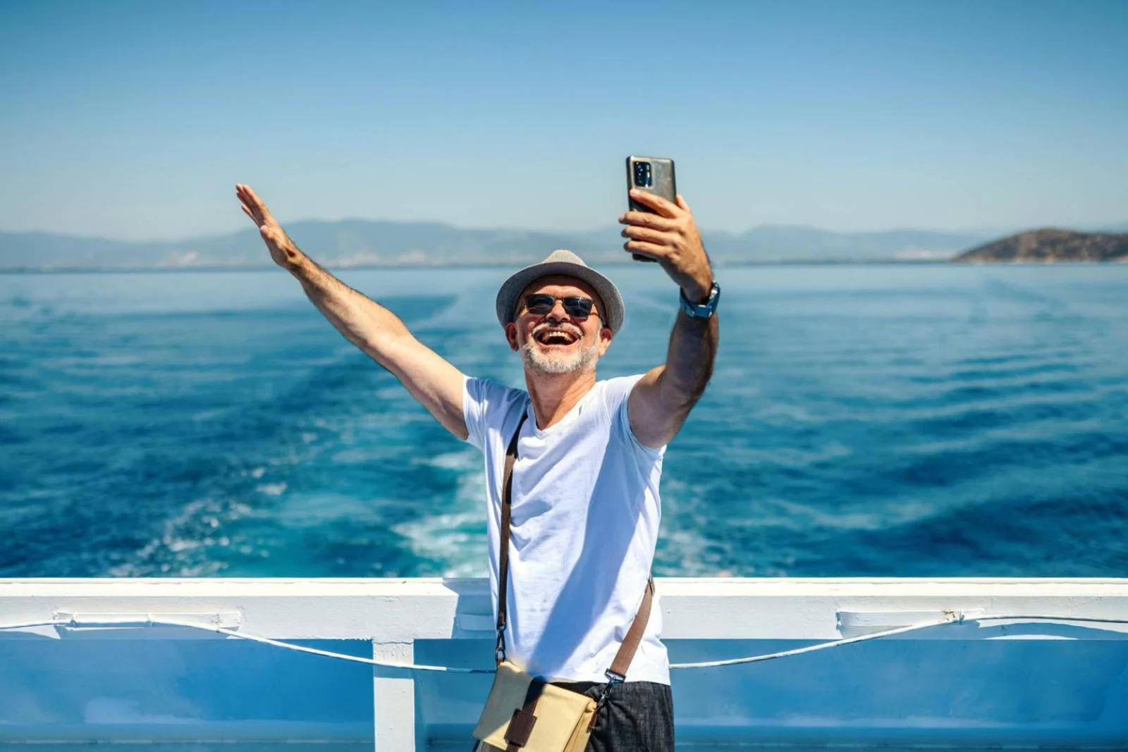 A mature man wearing a sunhat stands on the deck of a cruise ship, smiling and taking a selfie