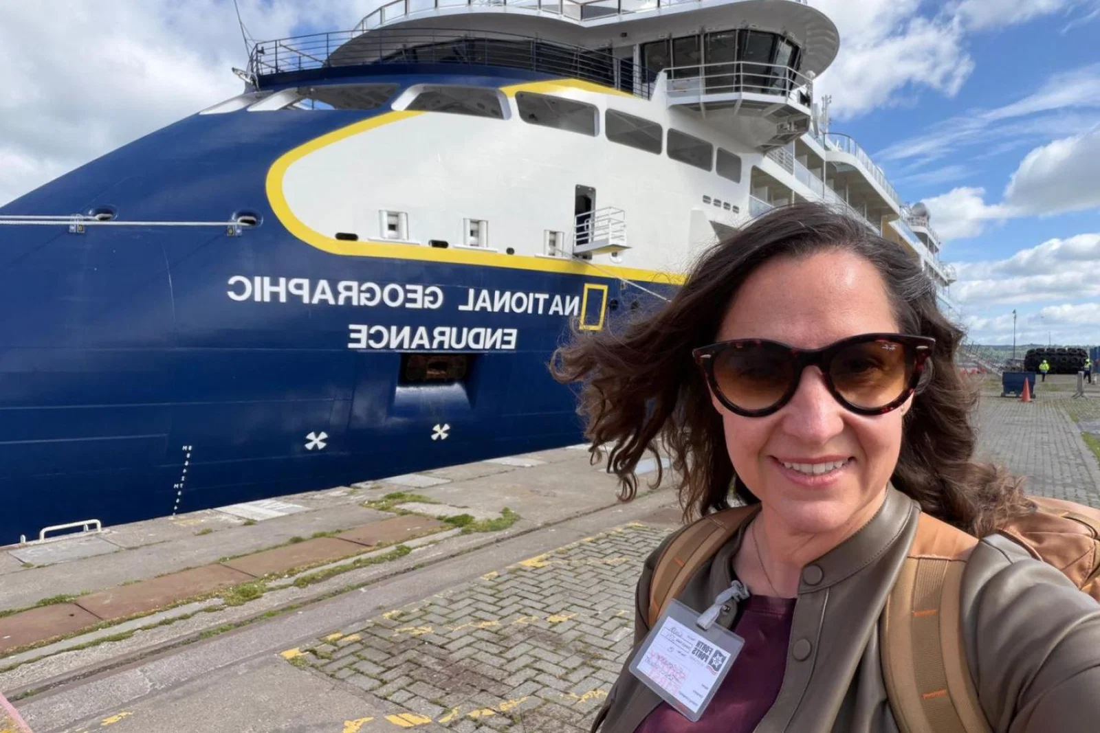 Woman with brown long hair and sunglasses smiling in front of blue and white ship 