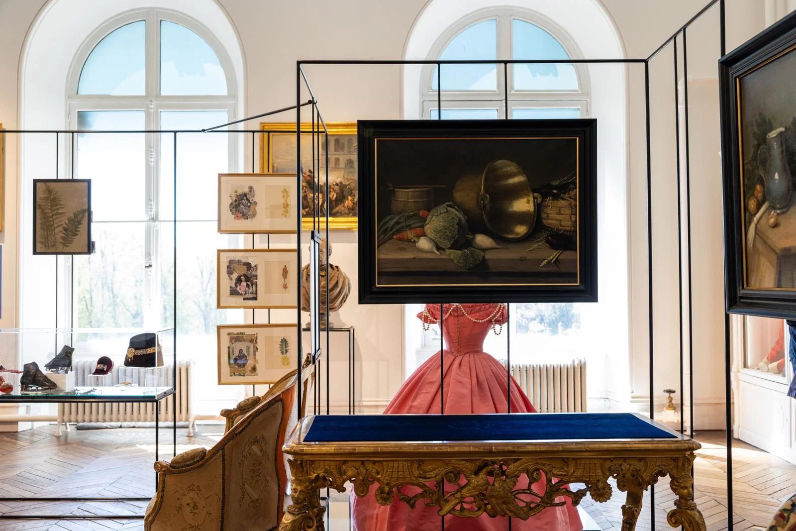 Photograph of an art gallery interior showcasing framed paintings, sketches, and a vintage gramophone displayed within glass cases. Central focus on a pink Victorian-style dress behind a glass case with ornate wooden table in foreground, highlighting historical and artistic exhibits.