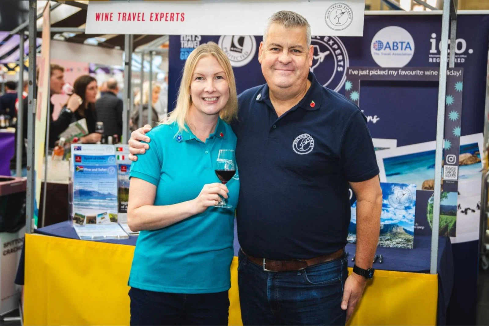 Blonde white woman and white man smiling at trade show, woman is holding glass of red wine 