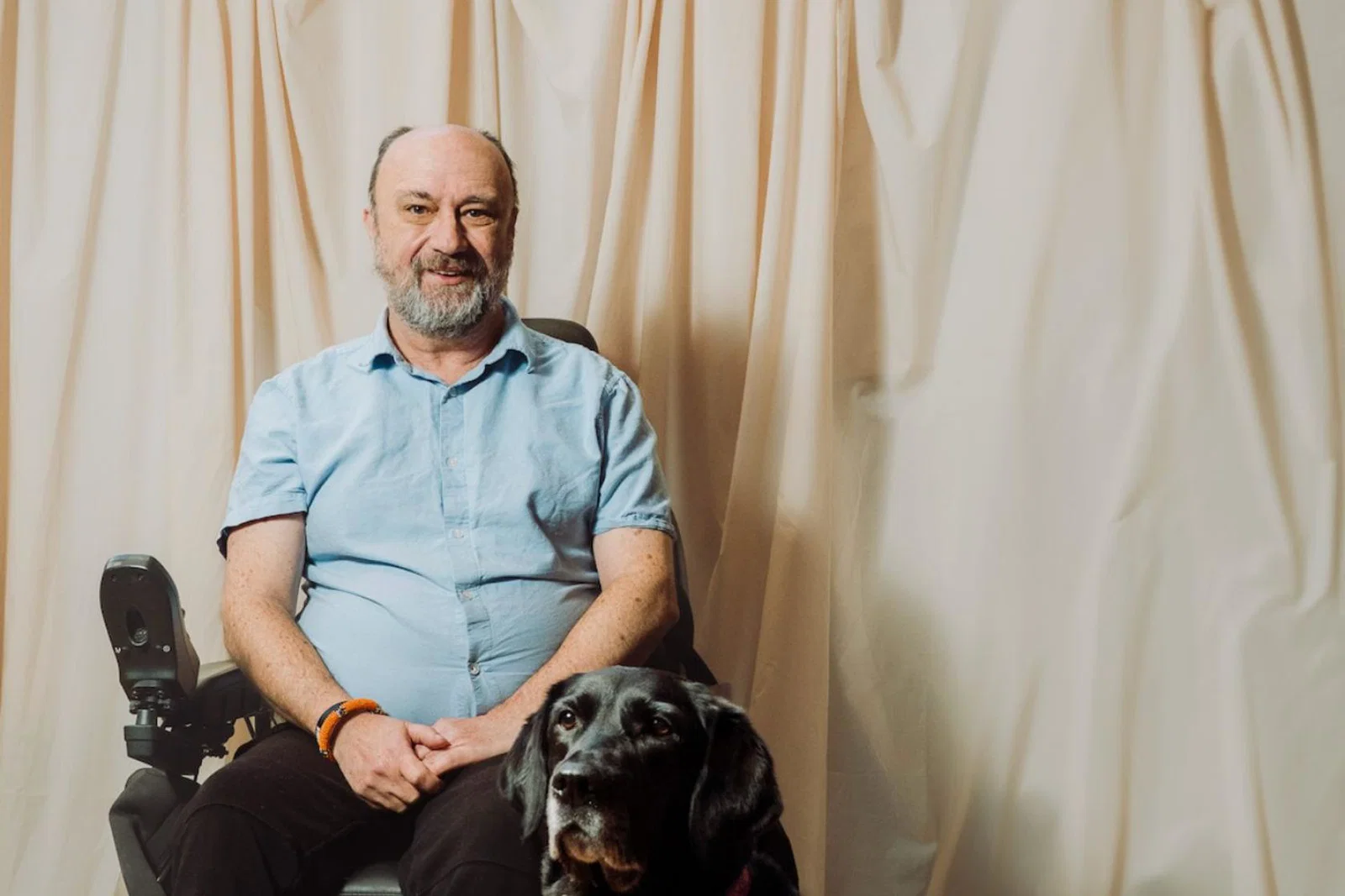 White man in his 50s, smiling in blue shirt, using wheelchair. A black dog is sitting to his right.  