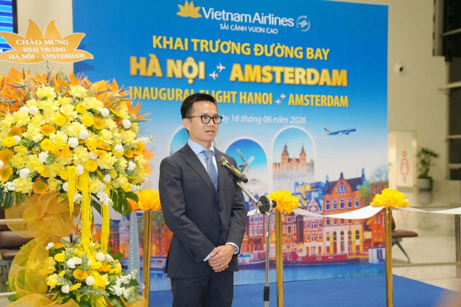 A man in a suit stands at a podium with microphones at a Vietnam Airlines event for the inaugural Hanoi to Amsterdam flight, with banners and yellow flowers in the background.