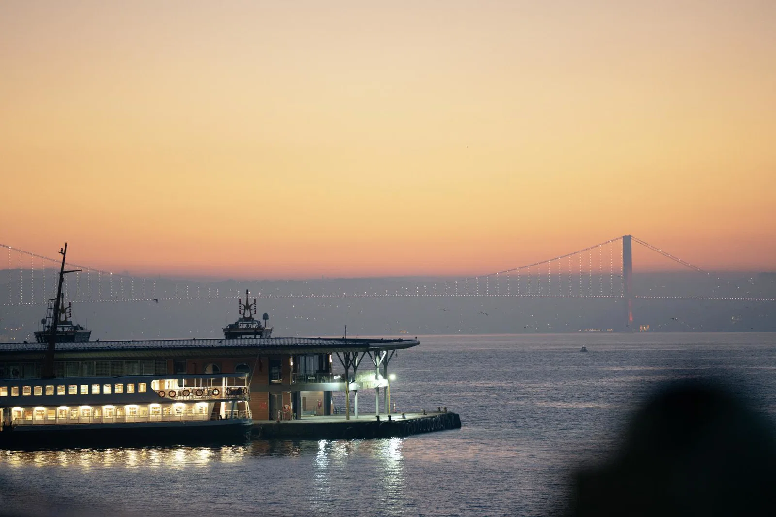 Istanbul’s waterfront and illuminated Bosphorus Bridge at sunset, with a ferry docked in the foreground.