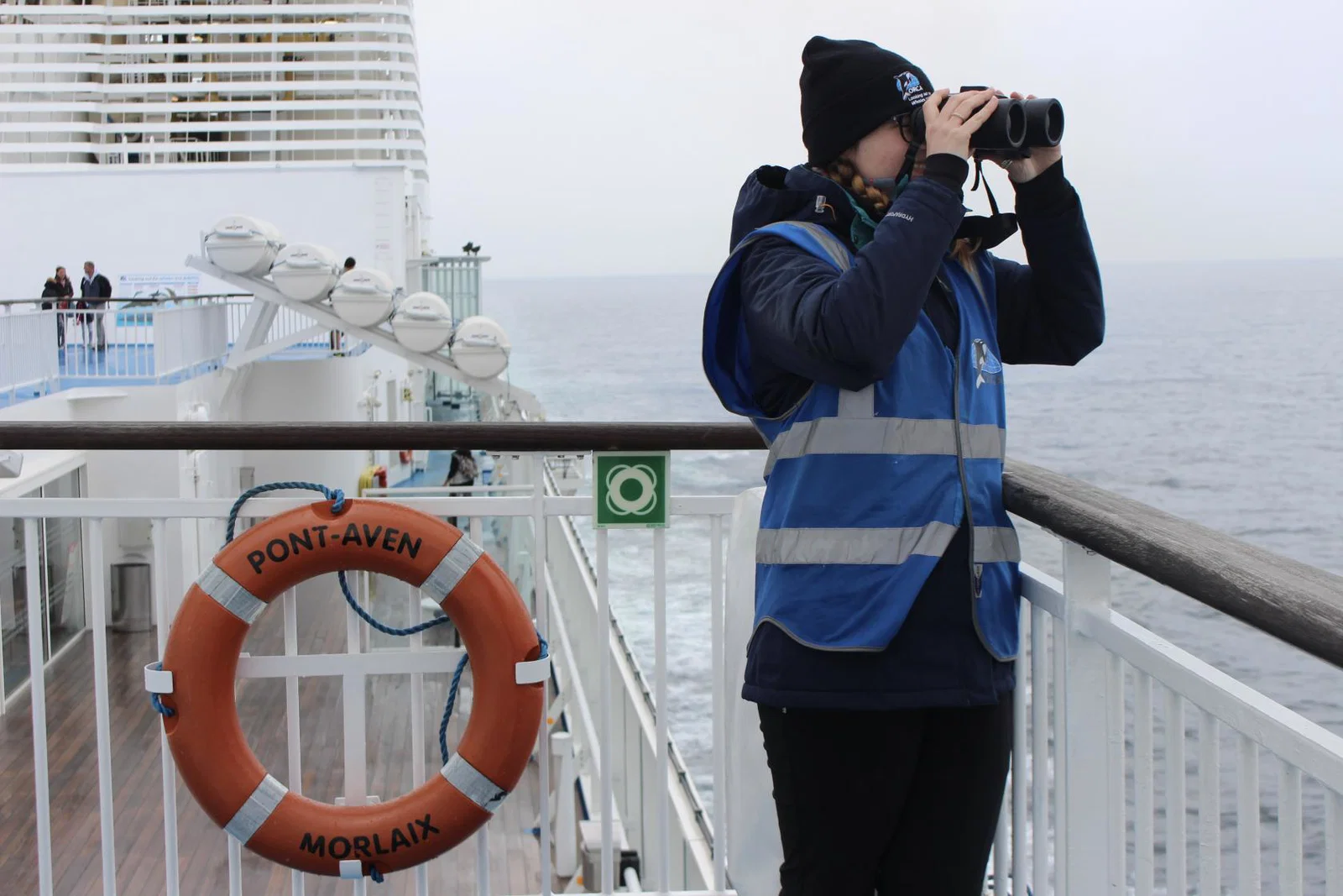 A wildlife surveyor from conservation charity ORCA stands on the deck of a ferry with binoculars looking out to sea in search of whales and dolphins.