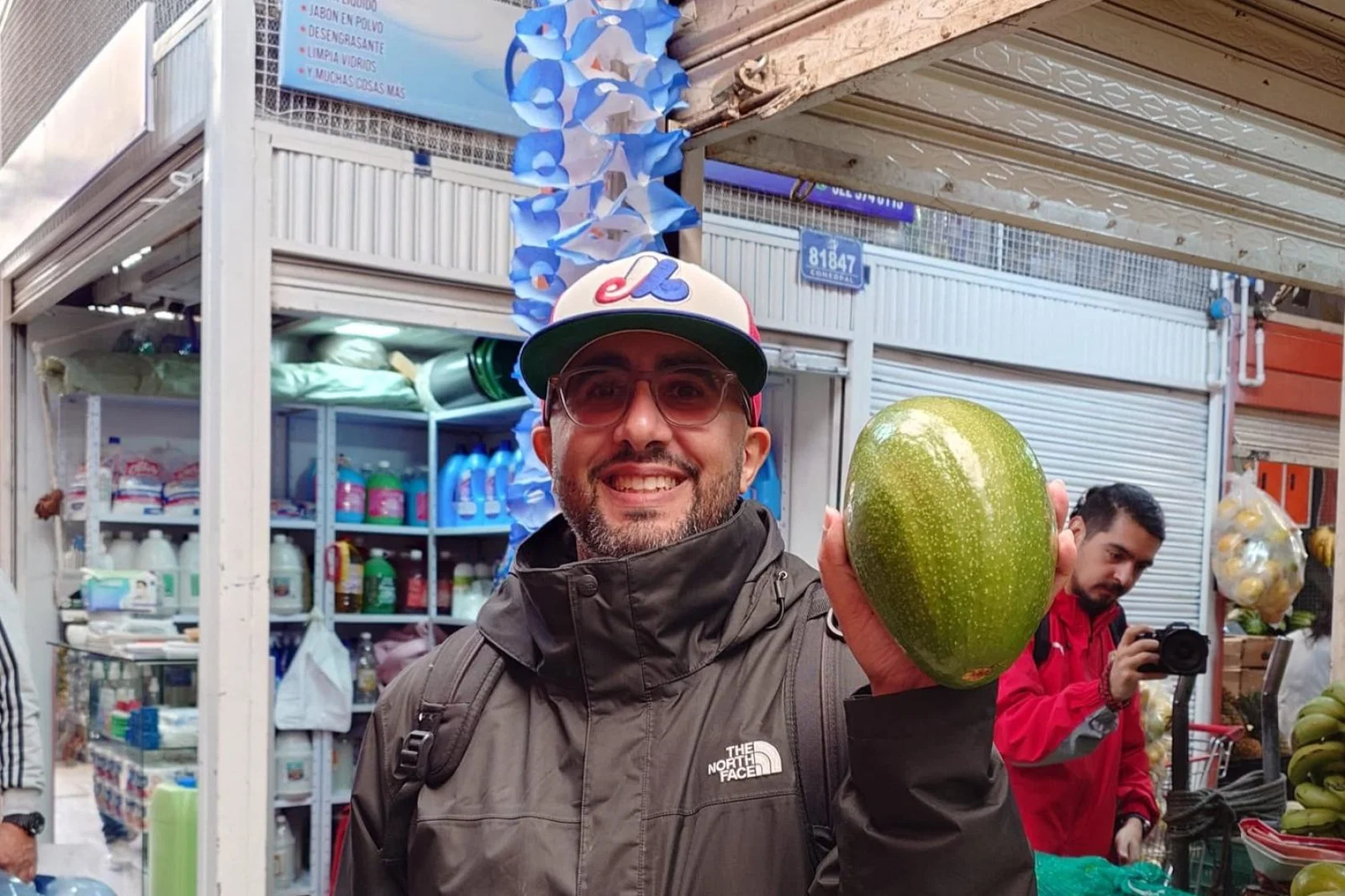 A smiling brown-skinned man with glasses and a baseball cap, holding up a large green fruit at a market