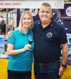 Blonde white woman and white man smiling at trade show, woman is holding glass of red wine 