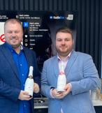 Two young white men standing in suits and holding champagne bottles