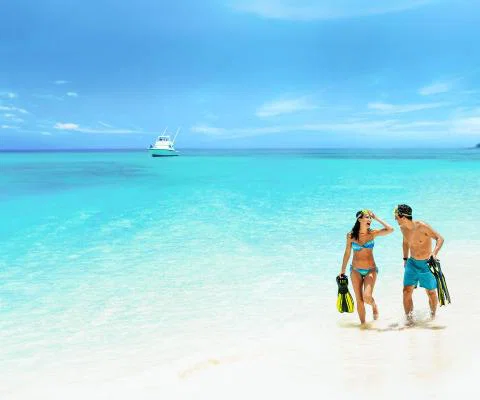 A young couple in swimwear walk along the white sand beach with their feet in the shallow of the blue sea