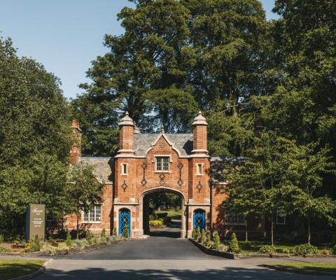 An ornate, 19th-century red-brick archway and gatehouse serves as the entrance to a luxury countryside estate, surrounded by mature green trees and manicured lawns under a clear blue sky. A modern hotel sign stands on the left edge of the driveway.