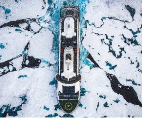 Aerial view of an expedition cruise ship navigating through fractured sea ice in polar waters, its dark hull cutting a path through a mosaic of white ice floes and dark blue-green water below.