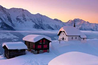 Three small wooden buildings in deep snow with mountains in background